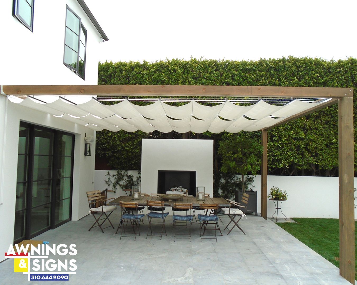 Pergola with fabric shade over outdoor dining area; white house, green hedge.
