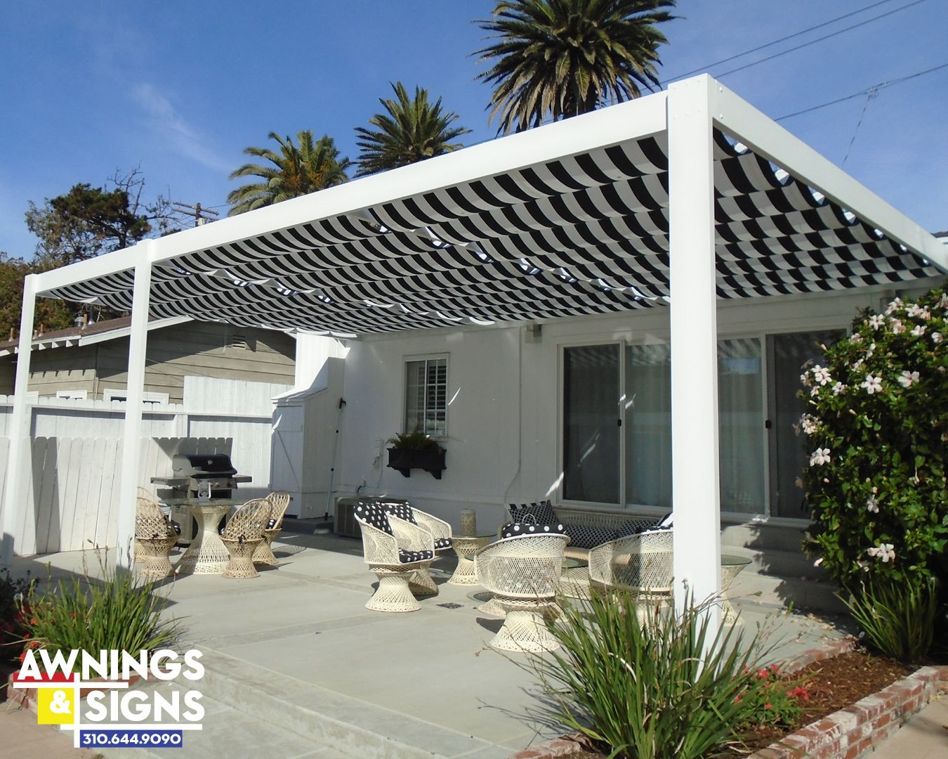 White pergola with black striped fabric, over a patio with furniture, white building in background.