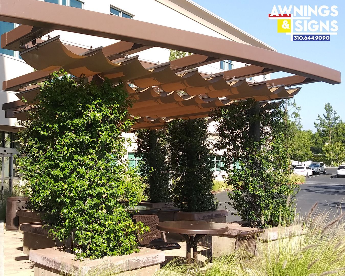 Brown pergola with tan fabric over seating area with shrubbery.