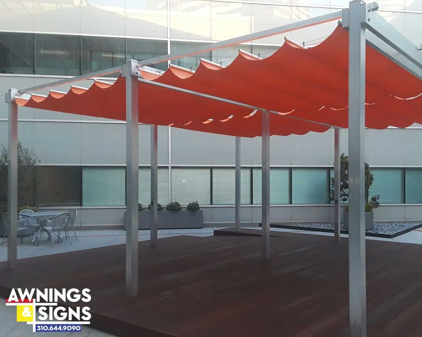 A modern pergola with an orange fabric canopy on a wooden deck. The structure has silver supports and a modern building in the background.
