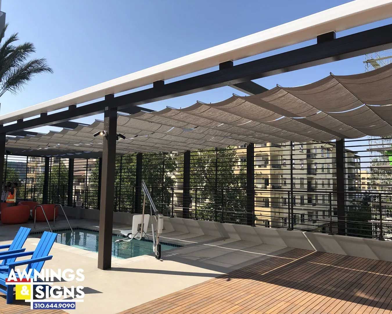 Outdoor patio with a pool and a retractable shade system over a seating area. Buildings and a blue sky are in the background.