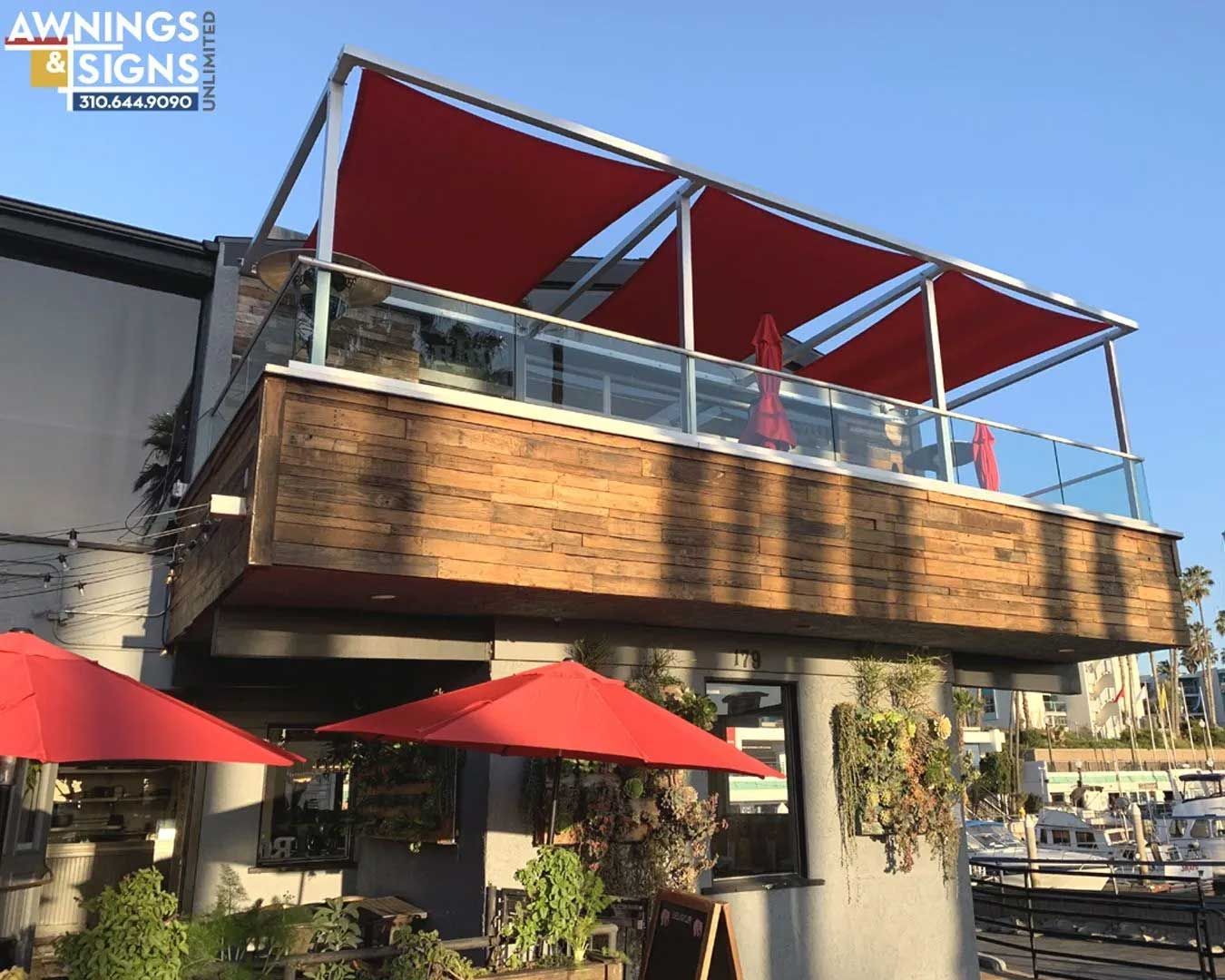 Red awnings shade a restaurant patio overlooking water. The modern building has a wooden facade and glass railings.