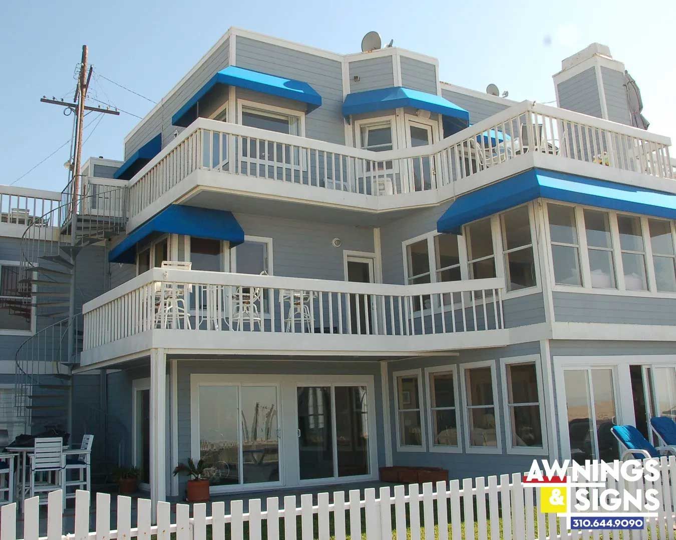 A multi-story, light-gray house with blue awnings over the windows and white railings, set on a sunny day.