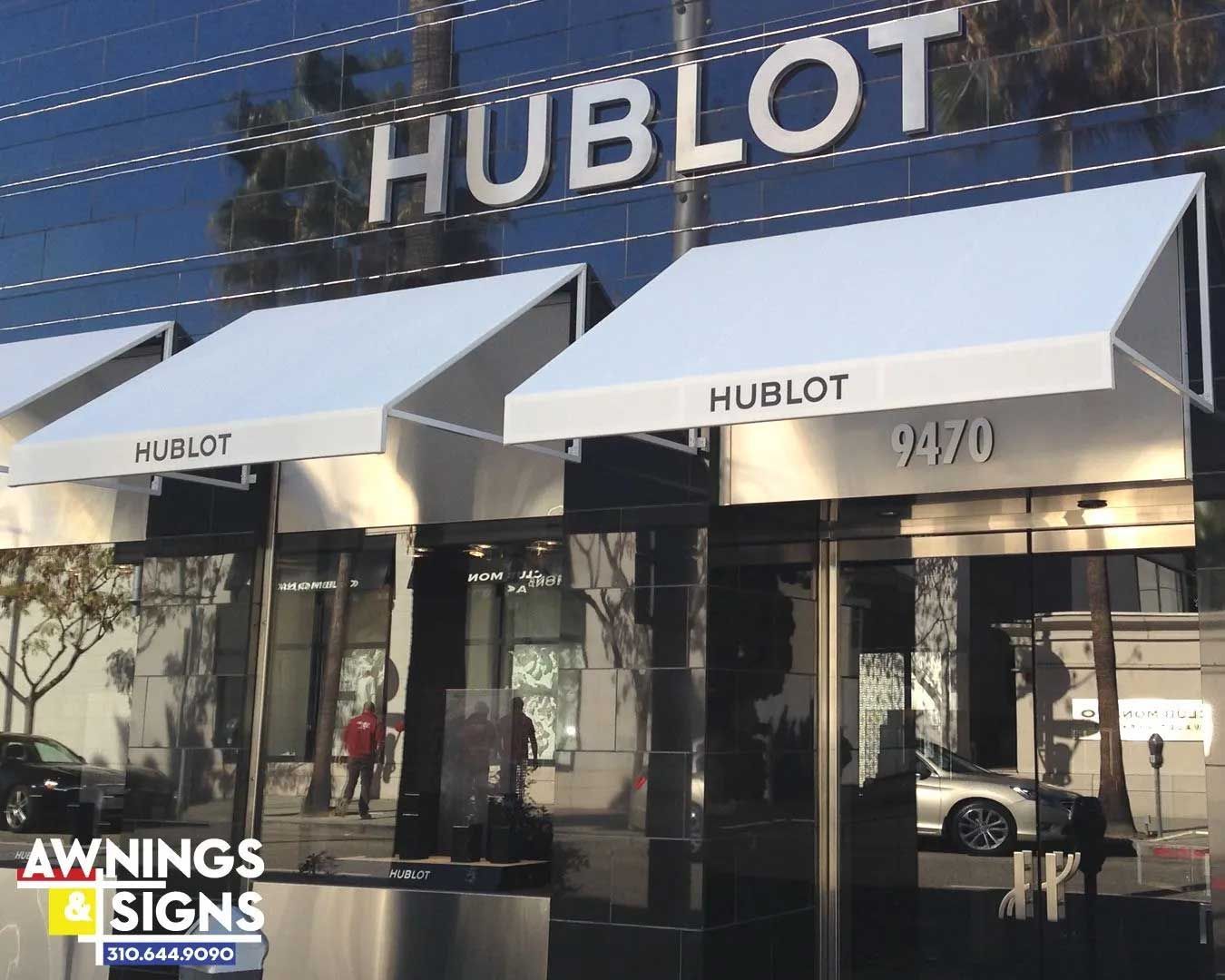 Exterior view of a Hublot store with white awnings, a reflective glass facade, and the address 