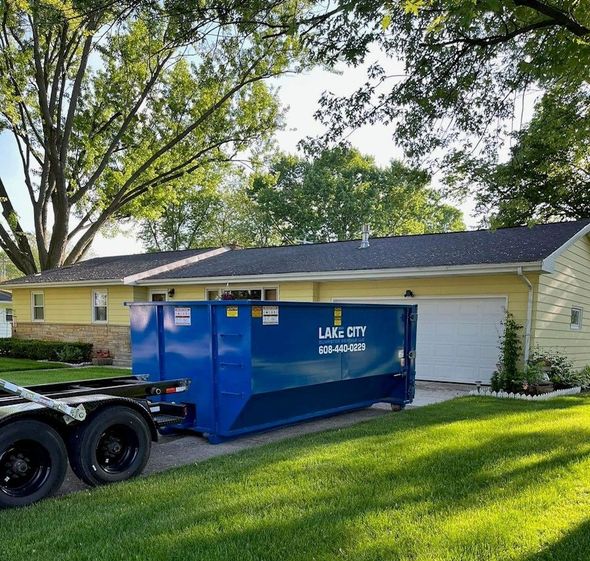 A blue dumpster from Lake City sits in the driveway of a yellow house.