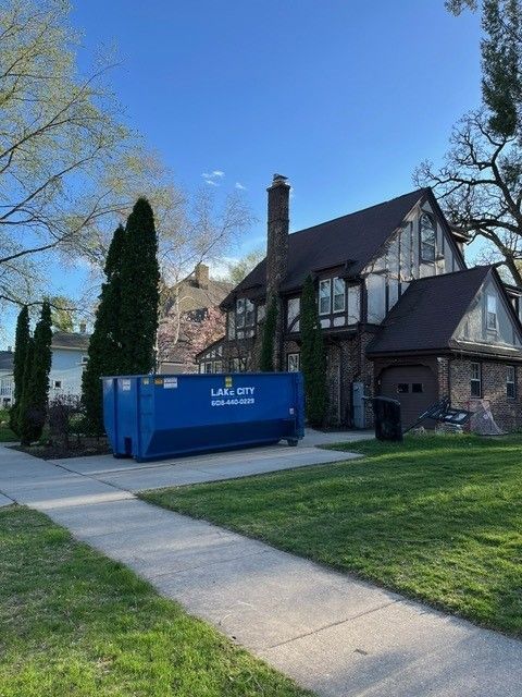 Blue dumpster sits in front of a Tudor-style house with a brick chimney and a green lawn. Sunny day.