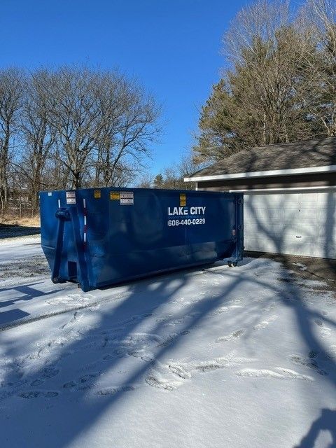 Blue dumpster in snowy driveway next to a garage under a bright blue sky.