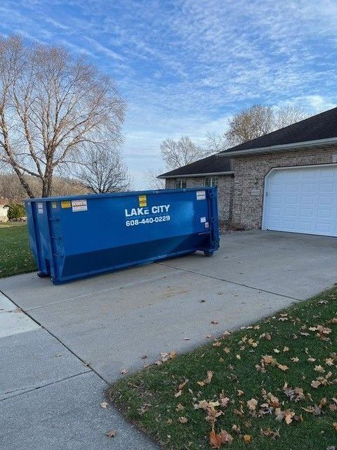 Blue dumpster on a driveway in front of a brick house and garage with bare trees and cloudy sky.