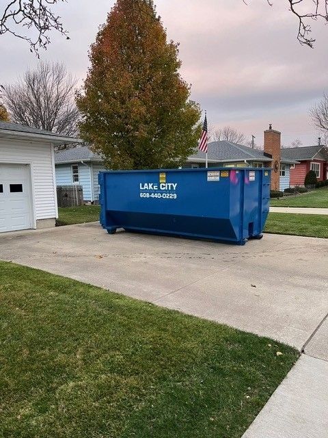 Blue dumpster in a driveway. House and lawn in the background, fall foliage, cloudy sky.