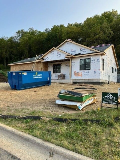 House under construction with blue dumpster, hillside in the background, and construction materials in the foreground.