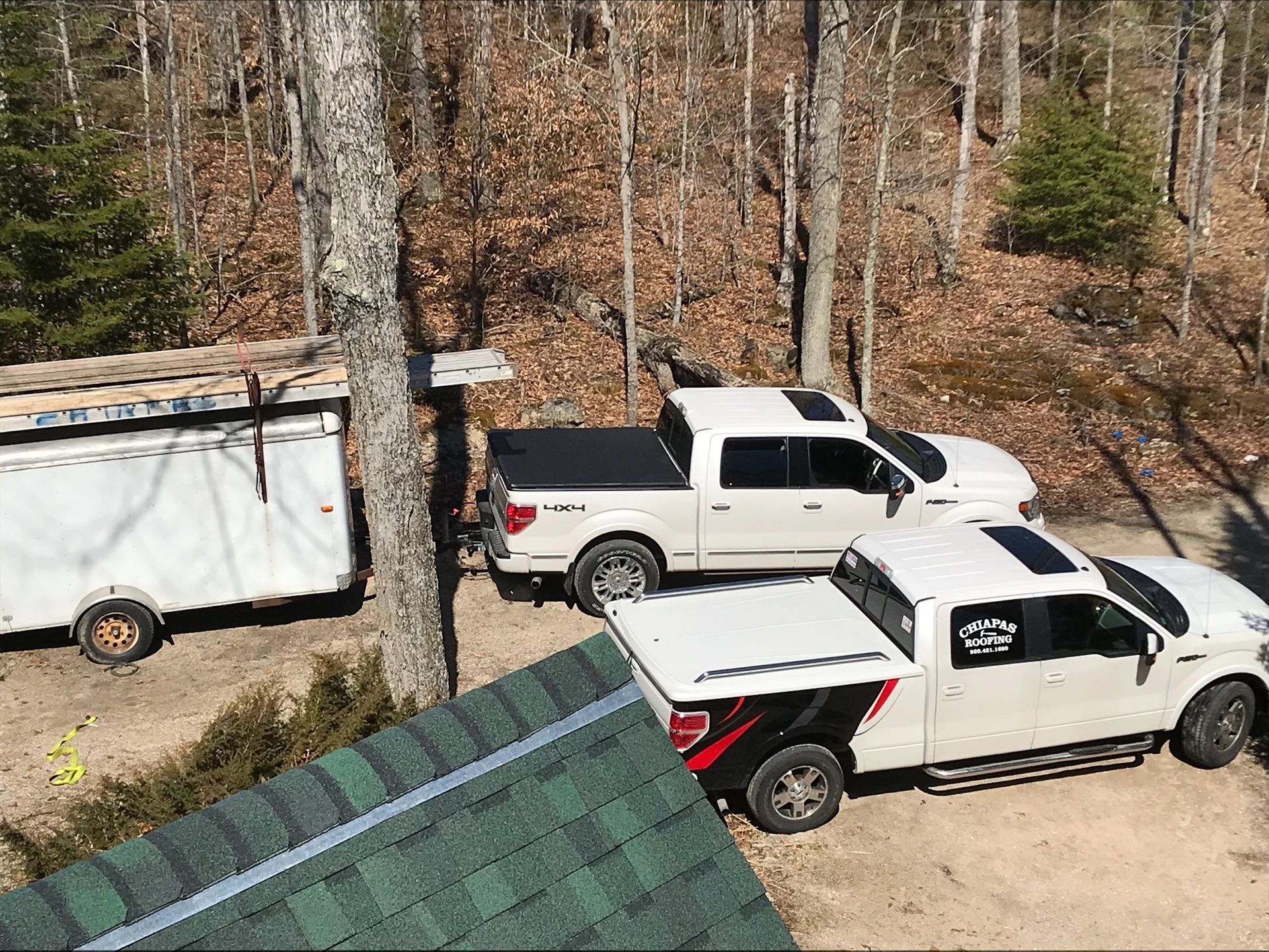Two white pickup trucks are parked next to a trailer in the woods.