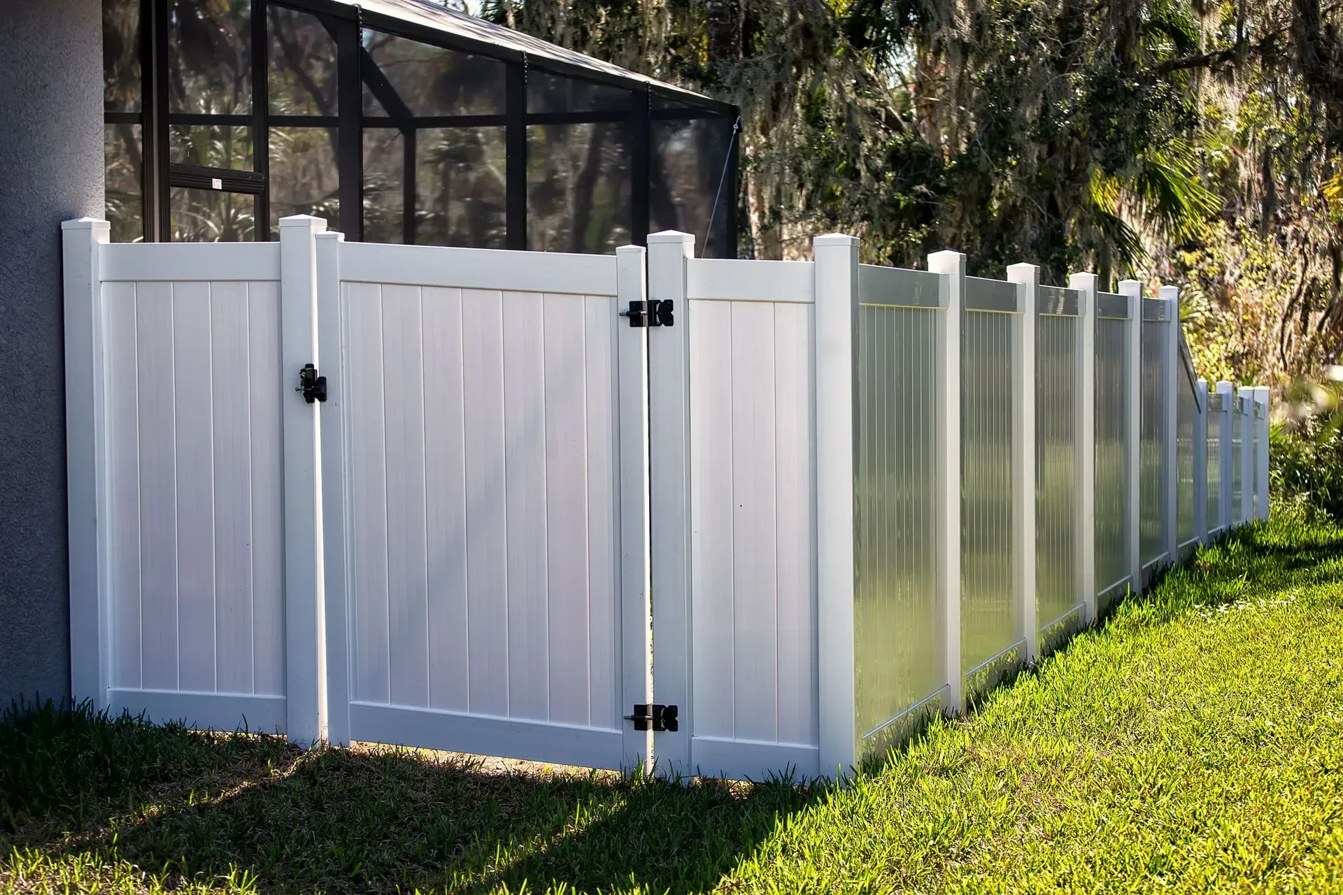 White vinyl fence with gate, bordering a green lawn near a screened porch and trees.