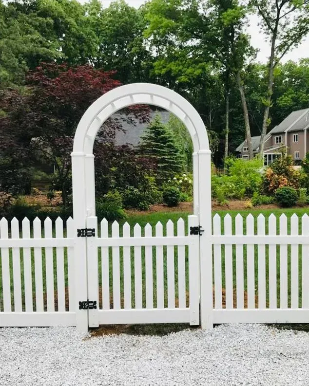White picket fence with archway gate leading to a garden with trees and a house in the background.