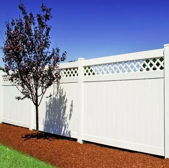 White vinyl fence with lattice top, red mulch bed, small tree, and blue sky.