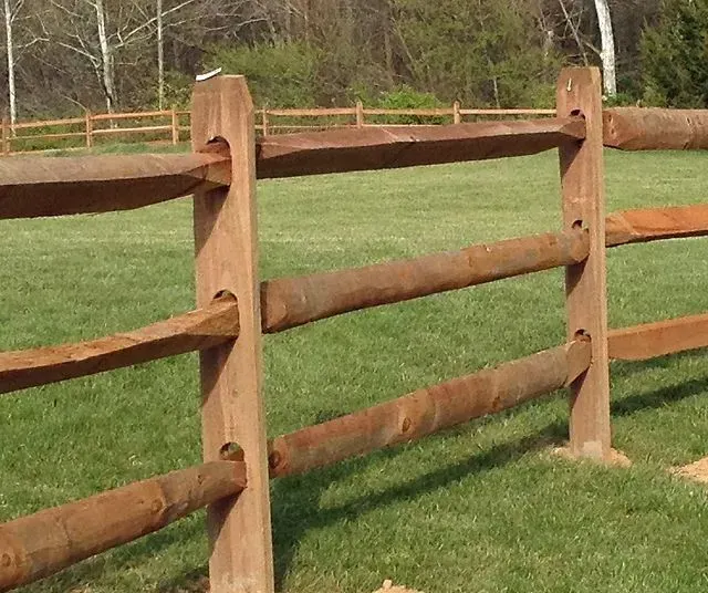 Wooden split-rail fence in a grassy field, with three horizontal rails connecting vertical posts.