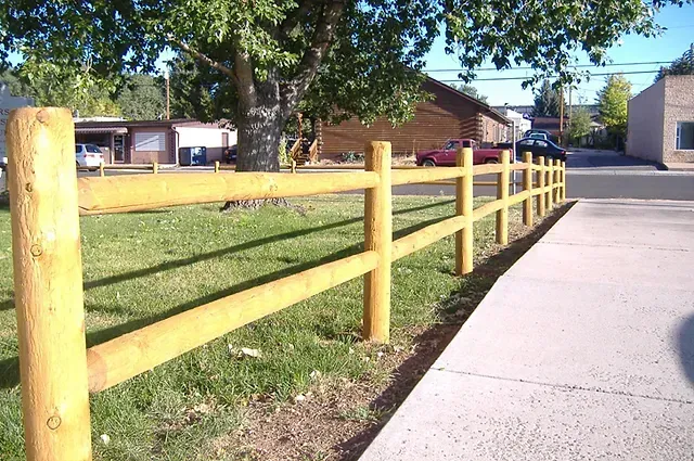Wooden split-rail fence alongside a sidewalk and grass, with a building and parked cars in the background.