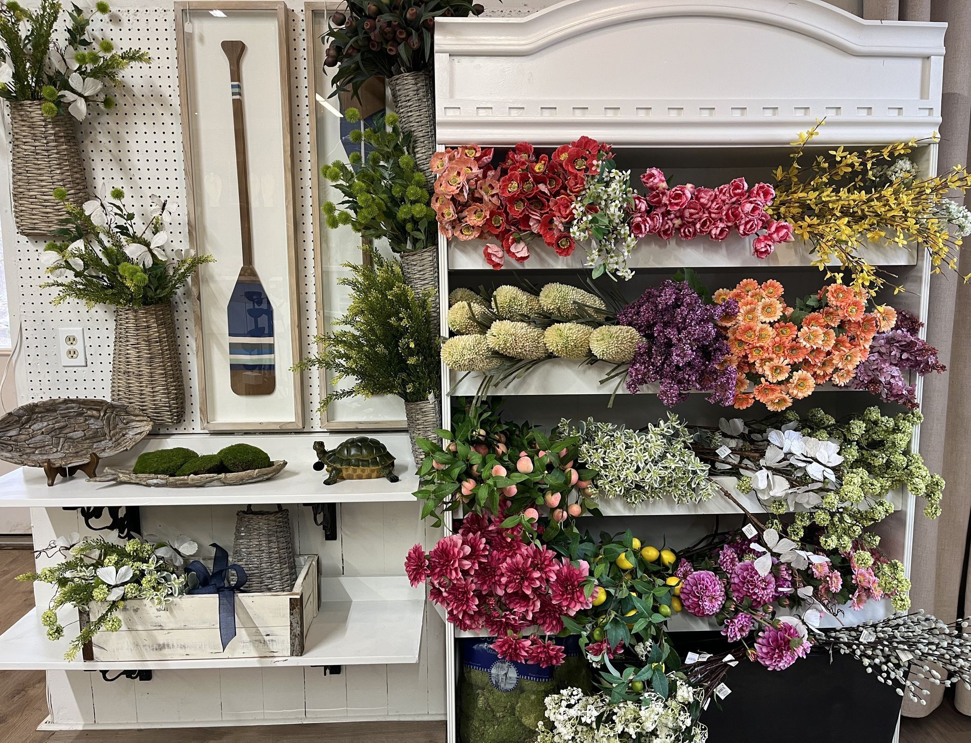 Display of colorful flowers and decorative items in a store. White shelves with various blooms and an oar.