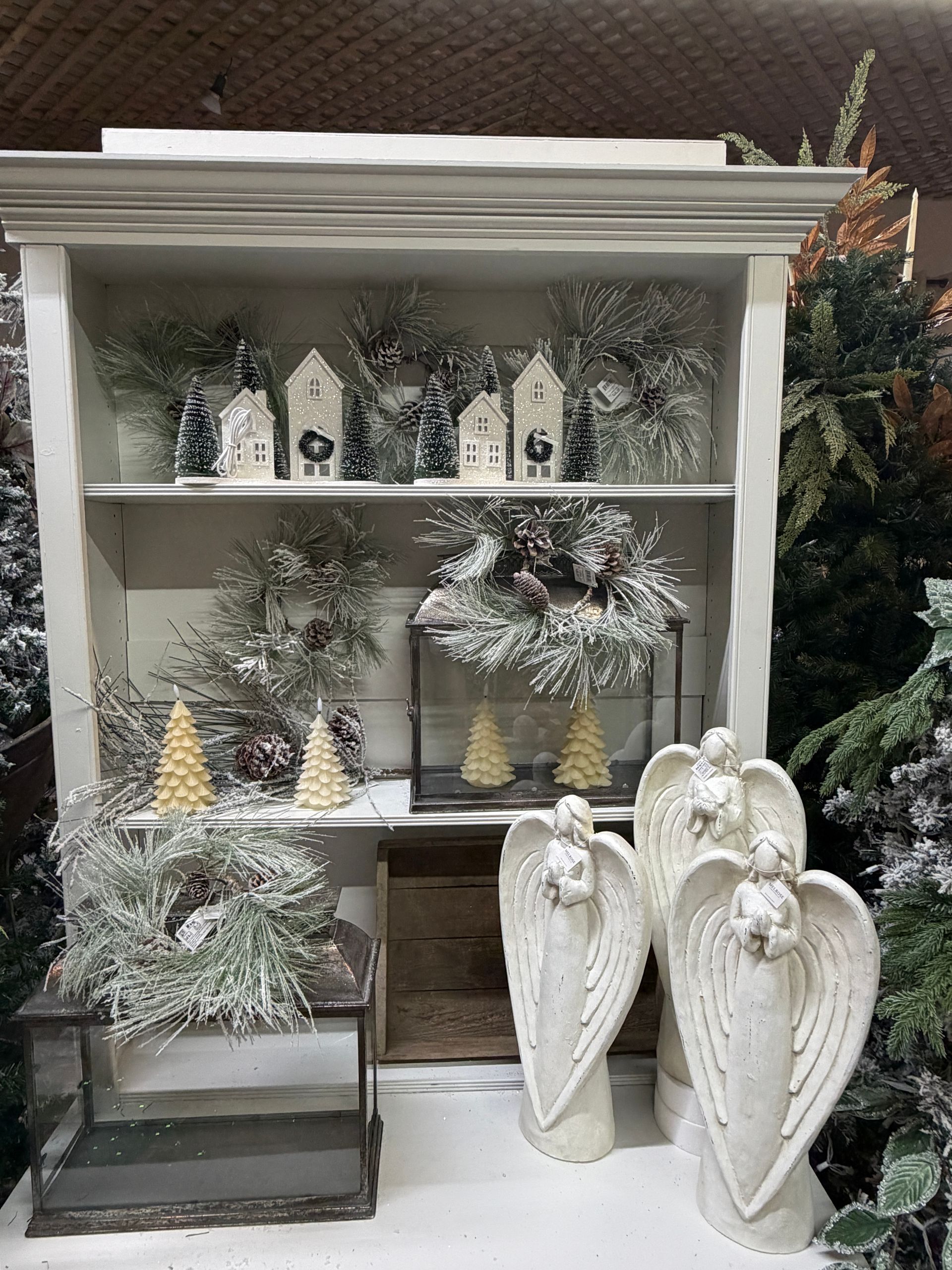 White display shelf with small winter decorations, miniature houses, frosted trees, and hanging angel wings.
