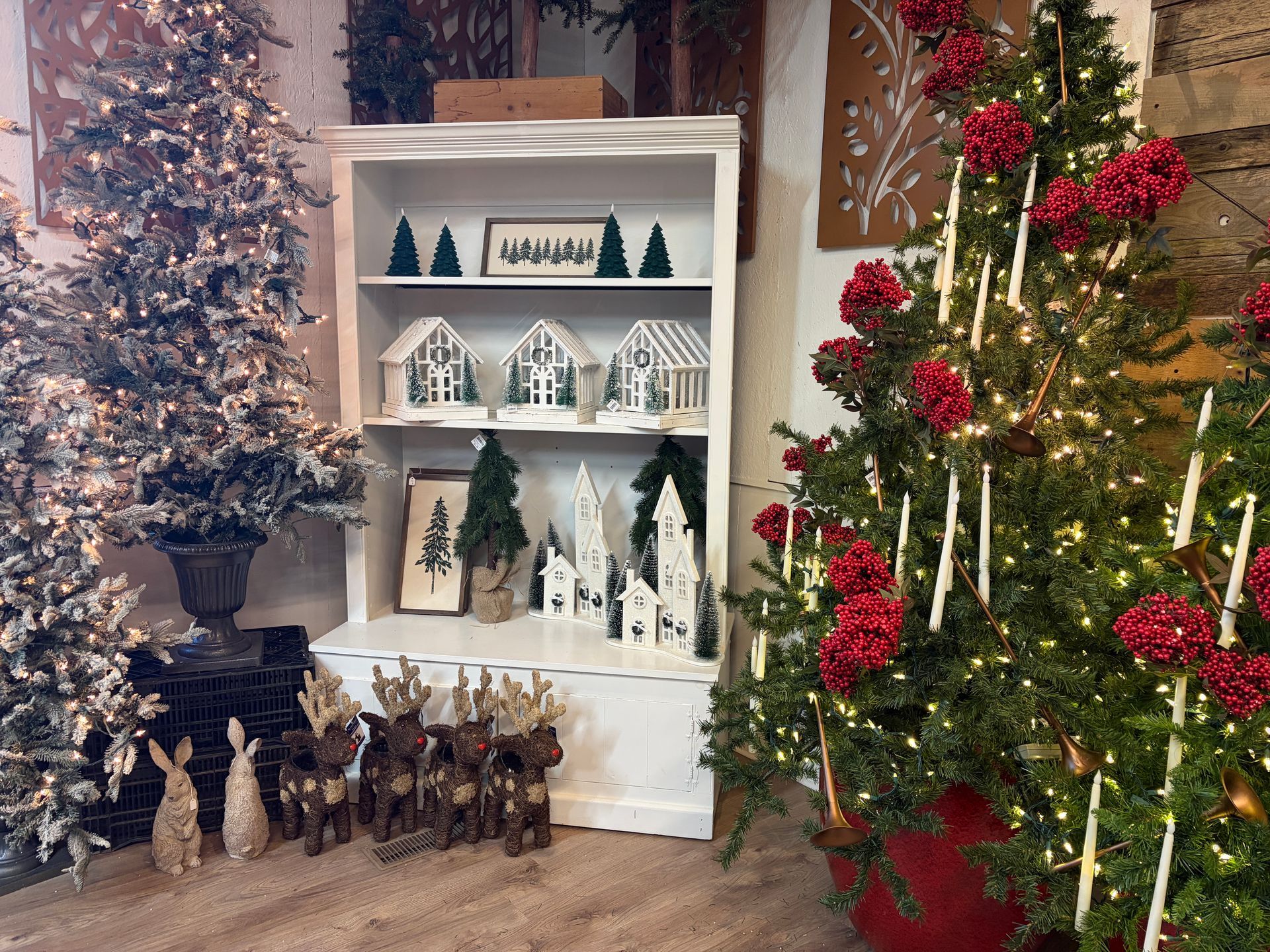 Holiday display with a lit Christmas tree, white shelf of mini houses, and frosted trees in a cozy room