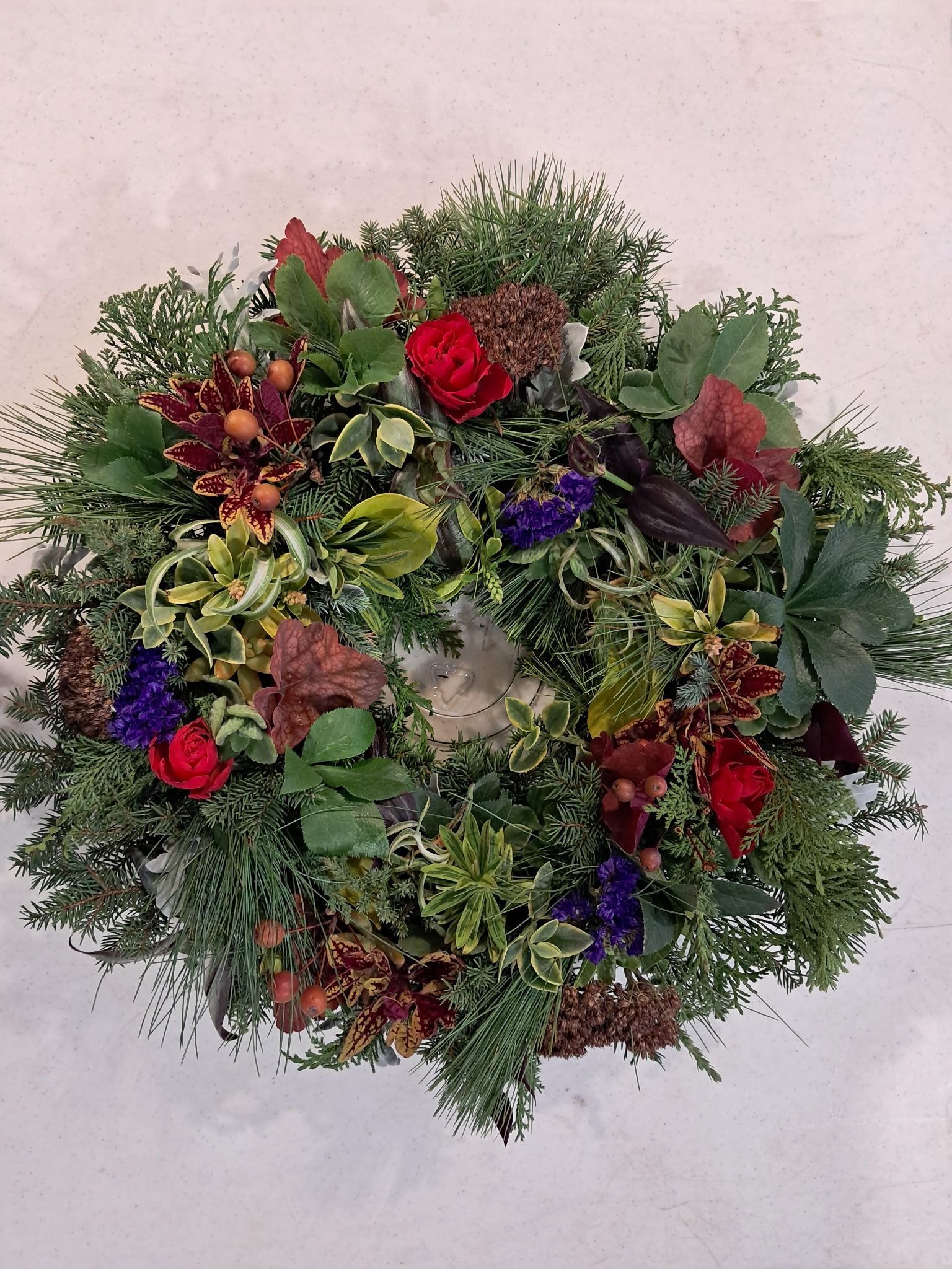 Wreath of evergreen and colorful flowers, including red roses and berries, on a light background.