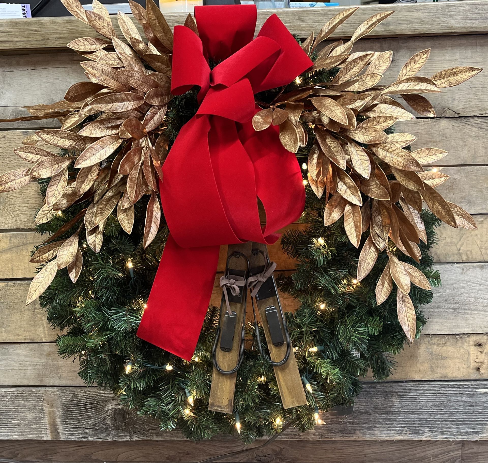 Christmas wreath with gold leaves, red bow, and small lights. Hanging against a wooden wall.