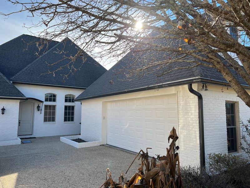 White house with black roof, garage, and tree in front; bright sunlight.