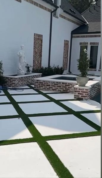 Backyard with white paved squares, green turf lines, brick walls, and a white building.