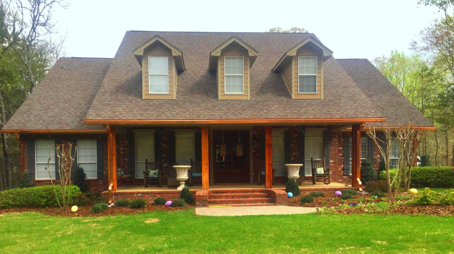 A brick house with a brown roof, porch, and green lawn.