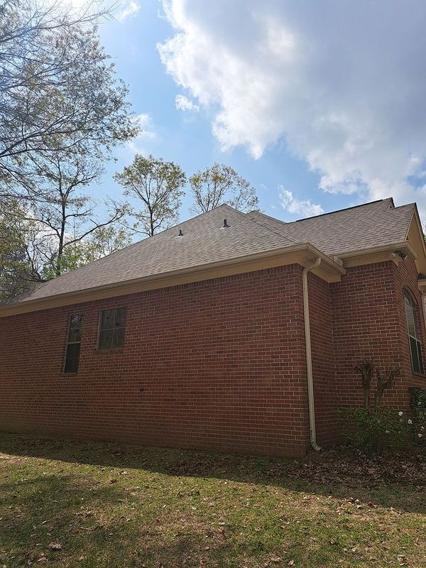 Brick house with brown roof, gutters, and windows. Trees and cloudy blue sky in background.