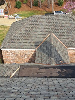View of a house roof with gray shingles and a brick wall. Trees and grass in the background.
