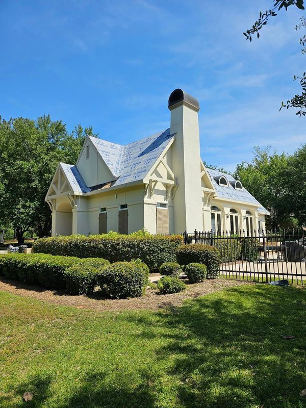 Cream-colored building with blue roof, chimney, and windows. Green hedges and grass in a sunny outdoor setting.