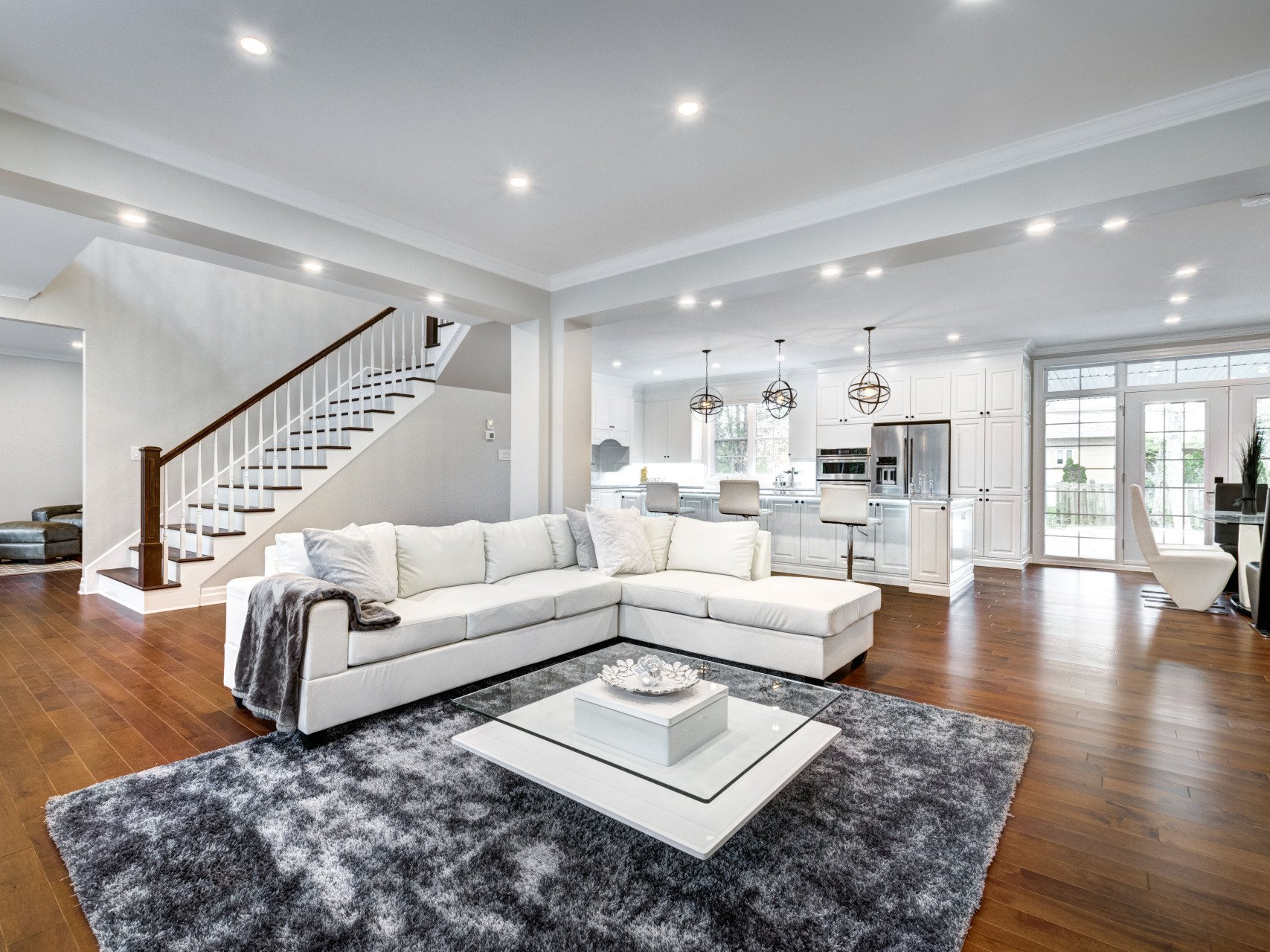 Spacious white living room with a sectional sofa, rug, stairs, and a kitchen visible in the background.