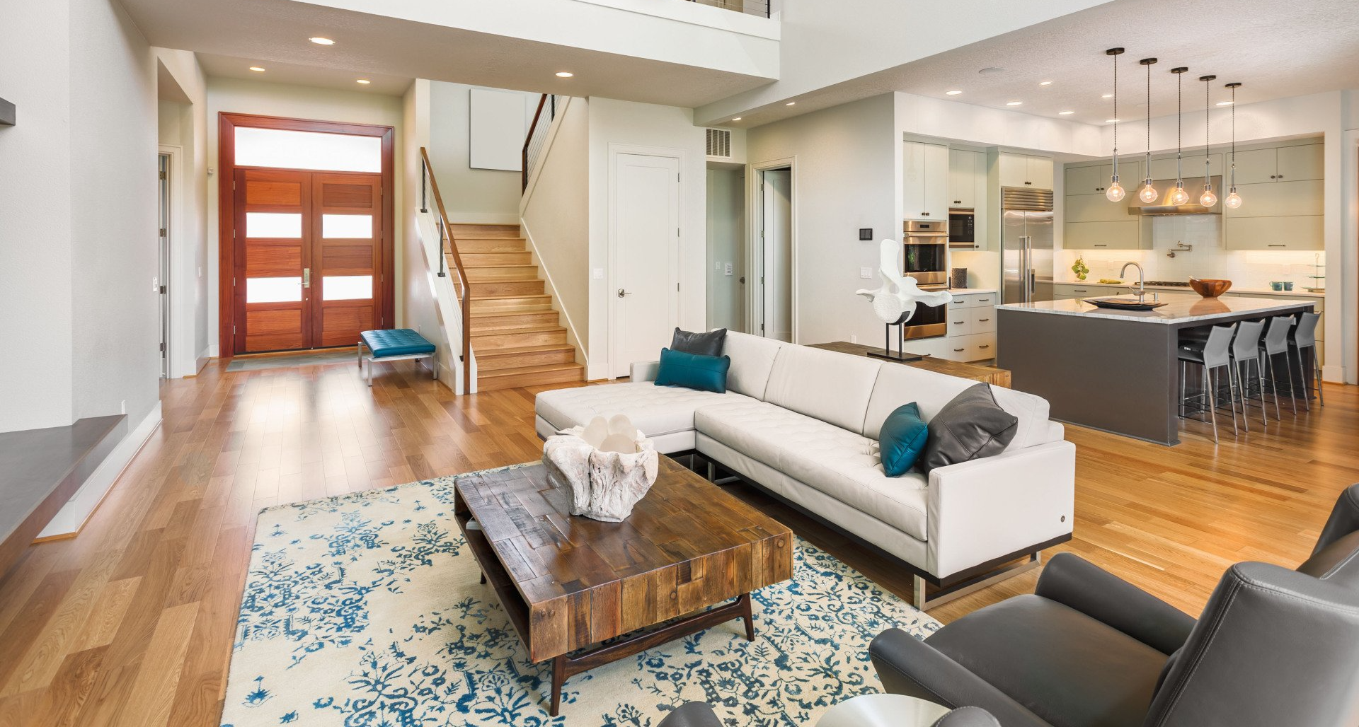 Modern living room with kitchen view, hardwood floors, white sectional sofa, and open staircase.