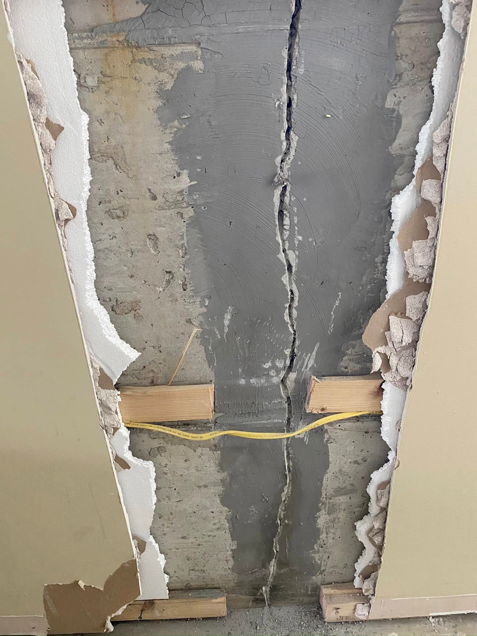 A section of drywall removed to reveal a large vertical crack in the concrete wall, with exposed wood framing and a yellow electrical wire.