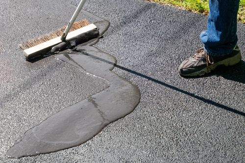 Worker spreading black sealant on pavement with a squeegee near a curbside crack