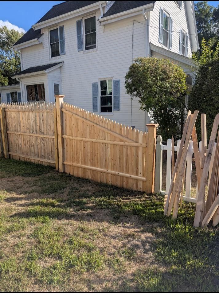 A wooden fence surrounds a white house. A white picket fence gate leads to the yard.