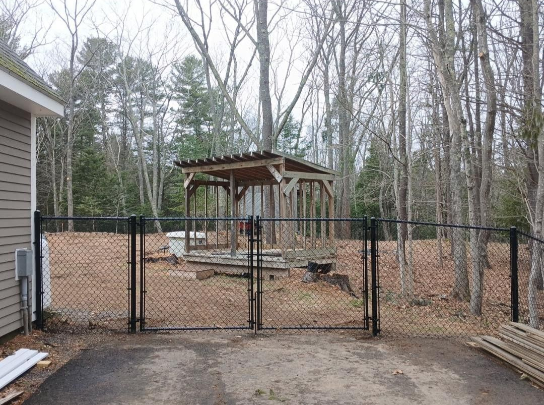 Black chain-link fence with double gate in front of a shed. The area behind the fence is a wooded area.