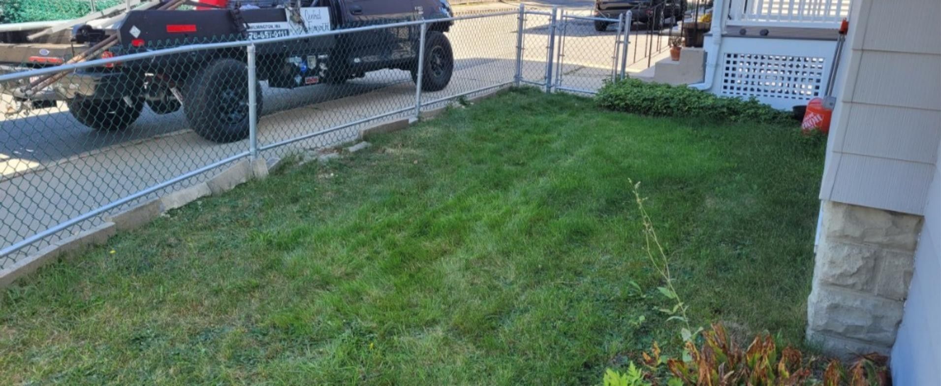 Lush green grass bordered by a chain link fence and a building. A truck is parked on the street.