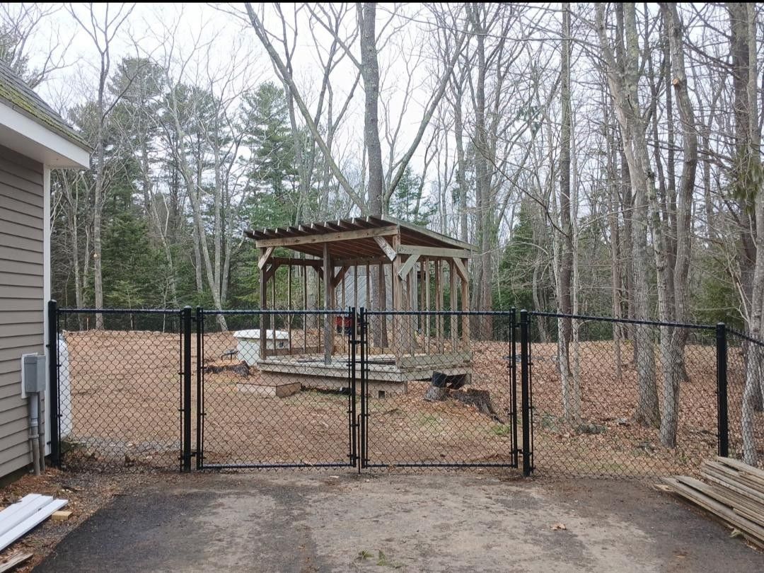 Black chain-link fence with gate in front of a wooded backyard, with a wooden gazebo structure in the center.