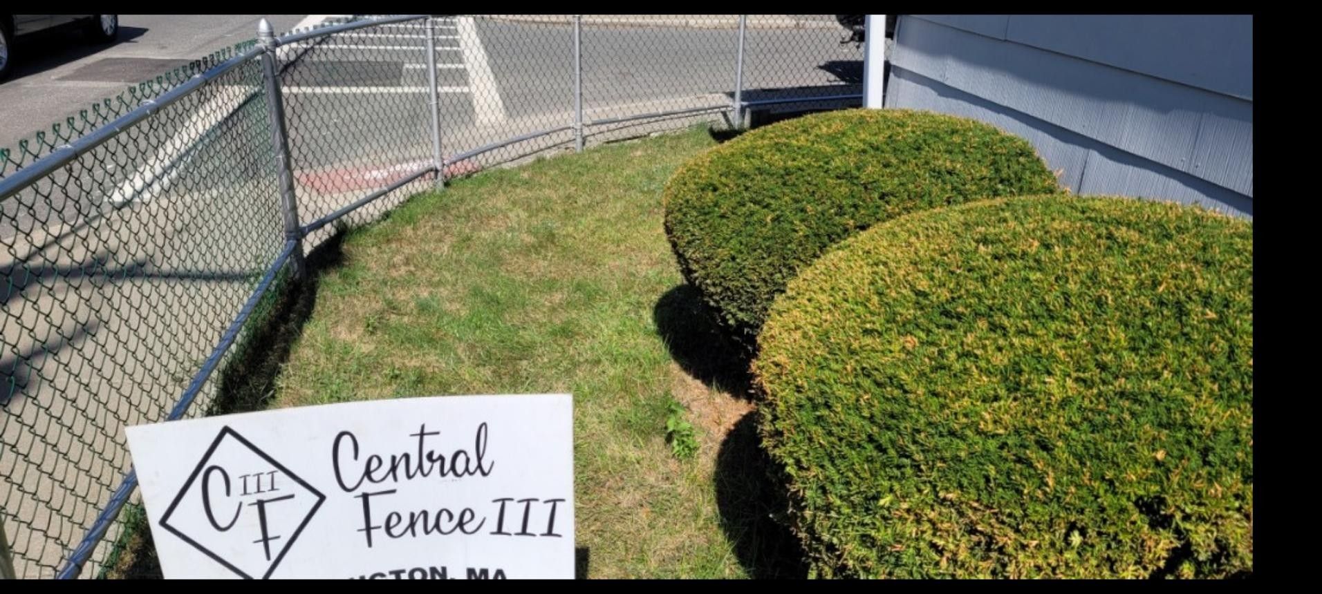A chain-link fence curves along a grassy area with three rounded, green bushes. A sign for Central Fence is in the foreground.