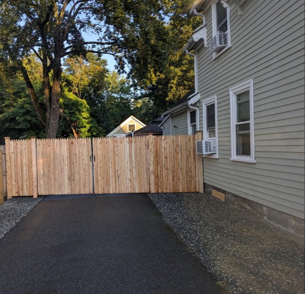 Wooden fence and driveway next to a light green house on a sunny day.