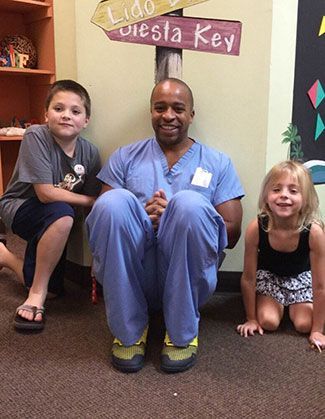 A doctor in blue scrubs sits on the floor with two children, smiling. They're near a beach-themed sign in a brightly colored room.