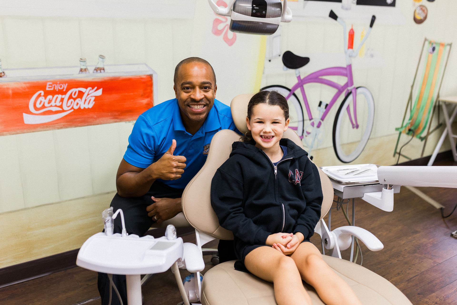 Smiling dentist with thumbs up and young girl in a dental chair, in a brightly painted dental office.