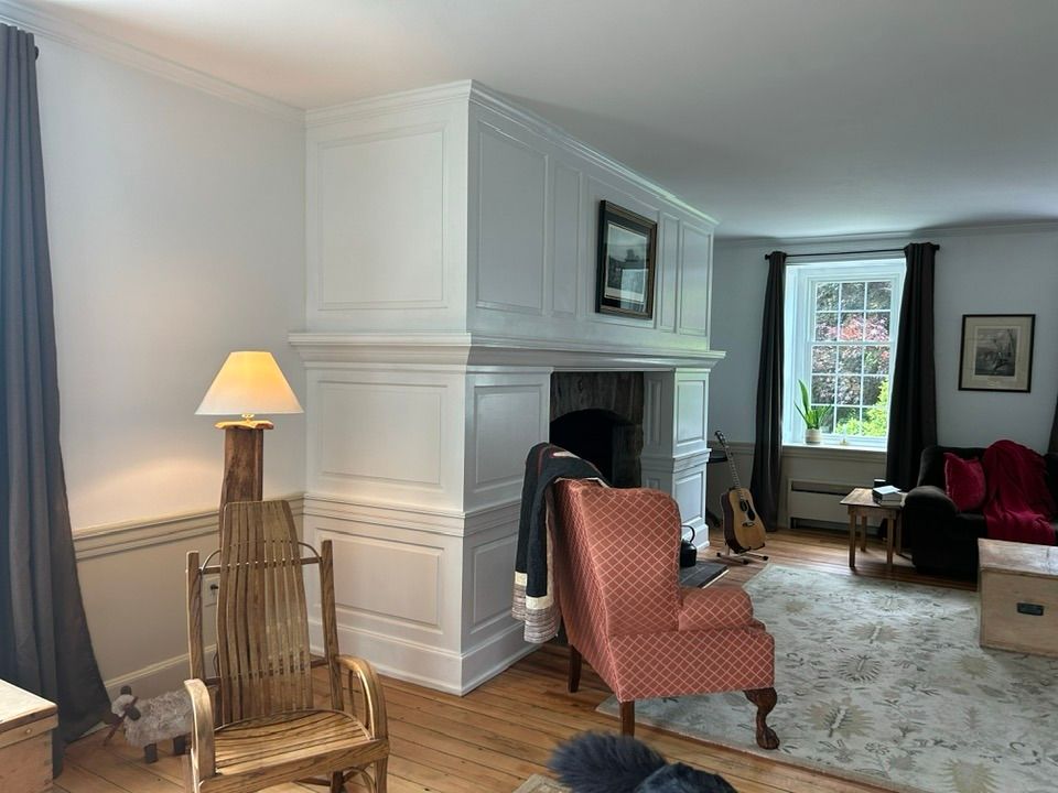 Living room with white fireplace, red chair, and wooden rocking chair. Window with dark curtains.