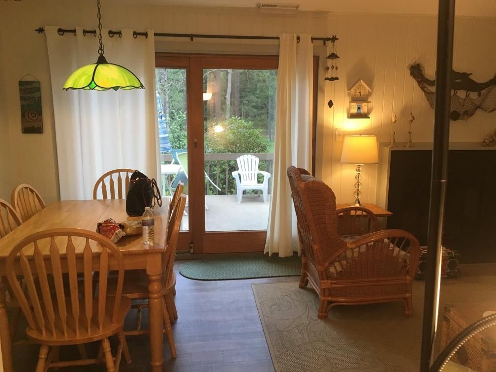 Dining room with wooden table and chairs, adjacent to a living area with a wicker chair, and glass doors opening to a deck.