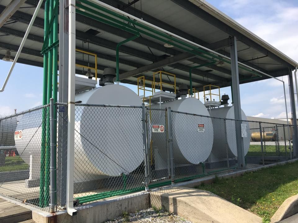 Fuel storage tanks behind a chain-link fence, under a metal roof, with green pipes.