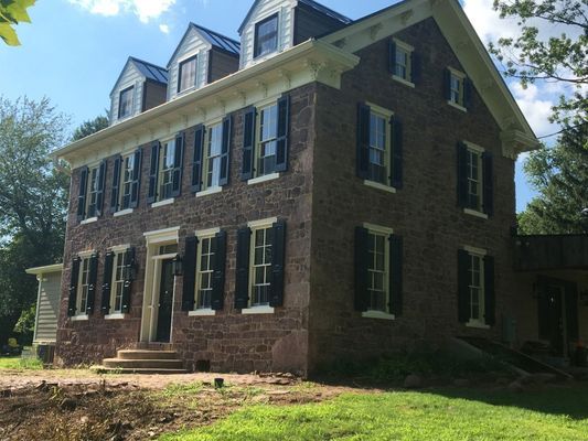 Three-story stone house with multiple windows, black shutters, and dormers under a blue sky.