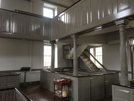 Interior view of a light-colored wooden church with a balcony and staircase, featuring windows and columns.
