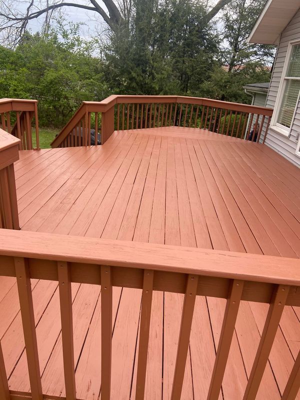 A freshly painted brown wooden deck with railings, next to a house and greenery.
