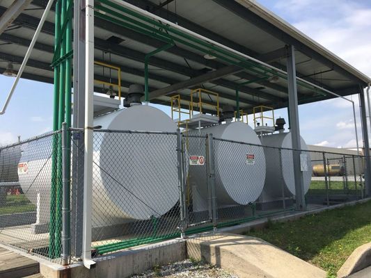 Three white fuel storage tanks behind a chain-link fence, under a metal roof, with green pipes and yellow railings.